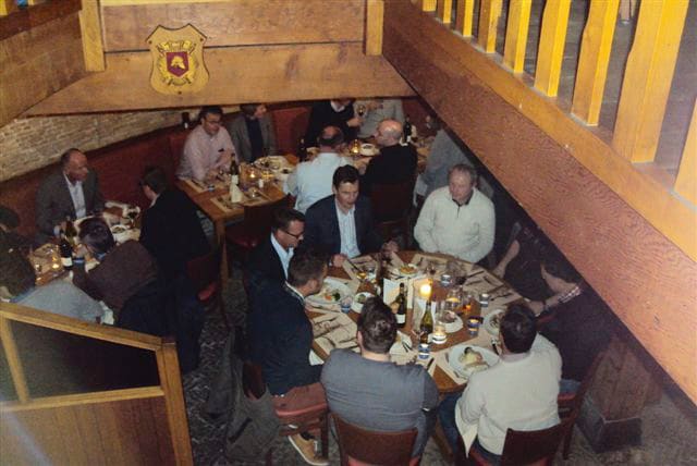 Een gezelschap aan lange tafels onder de houten balken van de molen, tijdens een besloten diner